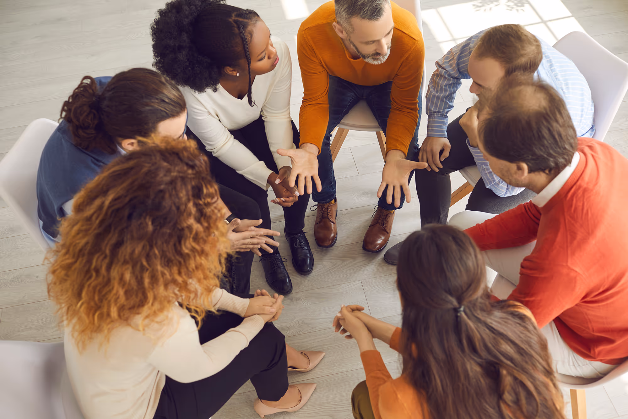A diverse group of people is sitting in a circle on chairs, engaged in a serious group discussion or support meeting. One man in an orange sweater is speaking with expressive hand gestures, while others listen attentively, suggesting a therapeutic or community support setting.