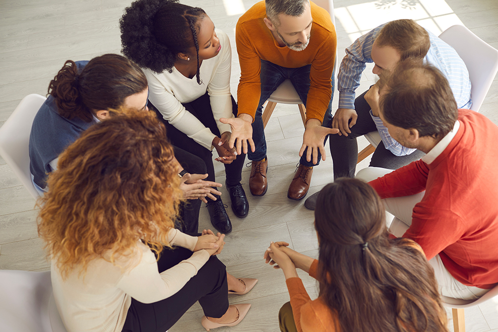 A diverse group of people is sitting in a circle on chairs, engaged in a serious group discussion or support meeting. One man in an orange sweater is speaking with expressive hand gestures, while others listen attentively, suggesting a therapeutic or community support setting.