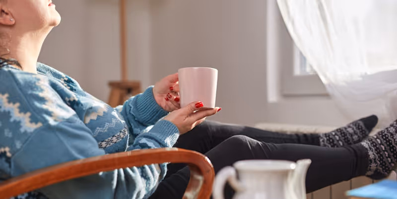 A woman sits in her personal dorm at a rehab centre, drinking tea and warming herself by the radiator, reflecting the quiet, restorative moments that are part of the healing process in heroin rehab.