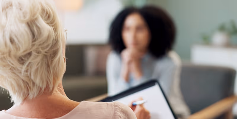 The image shows a counselling session between a therapist and a patient in a softly lit room. The camera focuses on the therapist’s hands holding a clipboard and pen, mid-consultation. The patient is seated opposite, slightly blurred and facing the therapist. The setting is warm and professional — with neutral tones, natural light, and minimal visual clutter. The body language appears open and attentive.