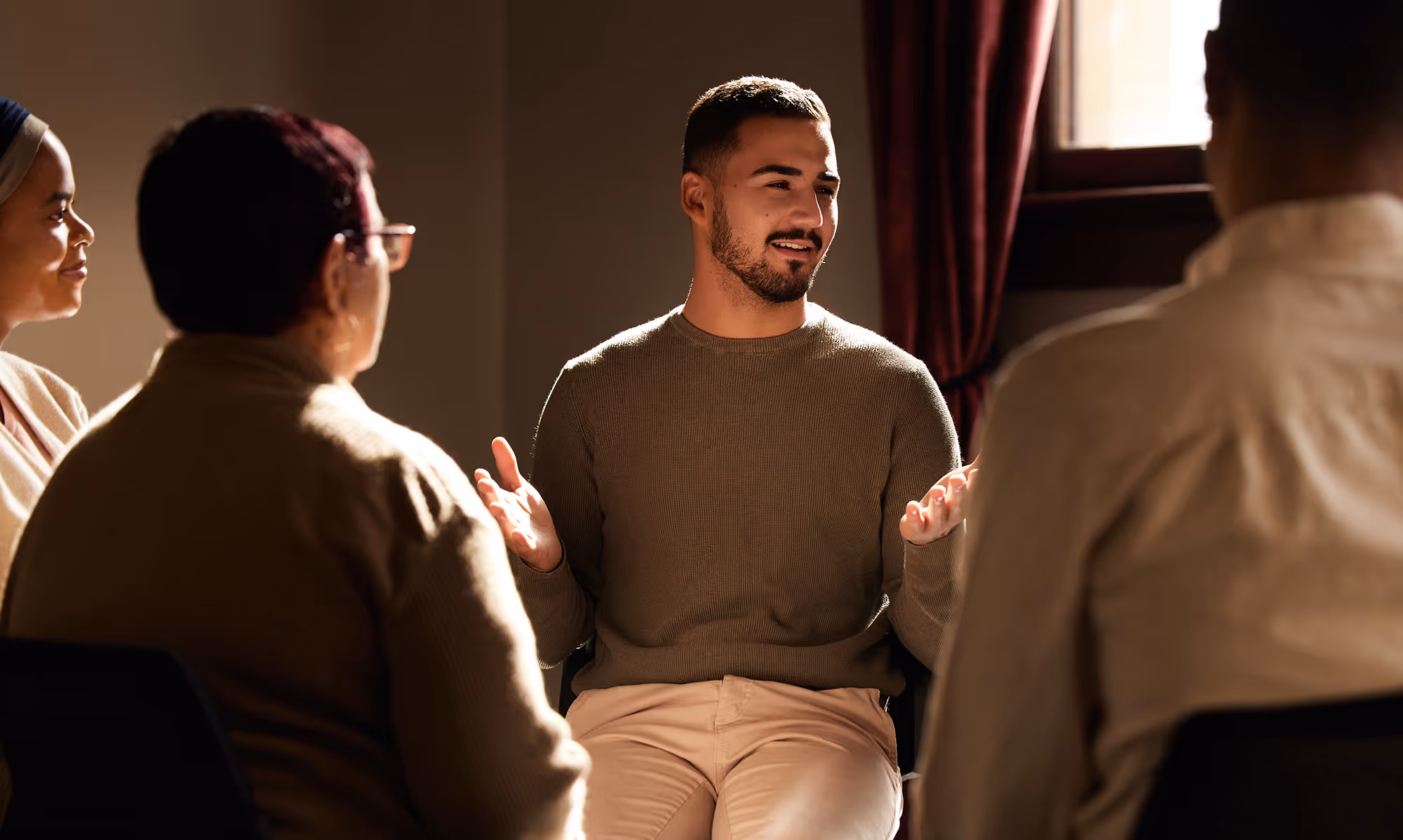 A group therapy session, in which a man is smiling and sharing with a mixed-gender group in a warm, safe therapy room.