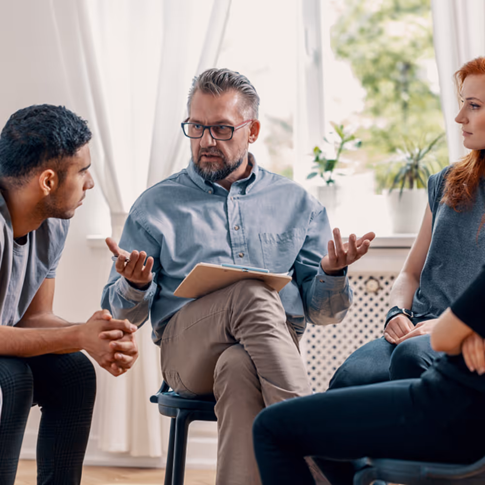 The same mixed-gender group therapy session on a sunny day. The group is sitting by an open window, but this time the therapist is sharing insights with the group.