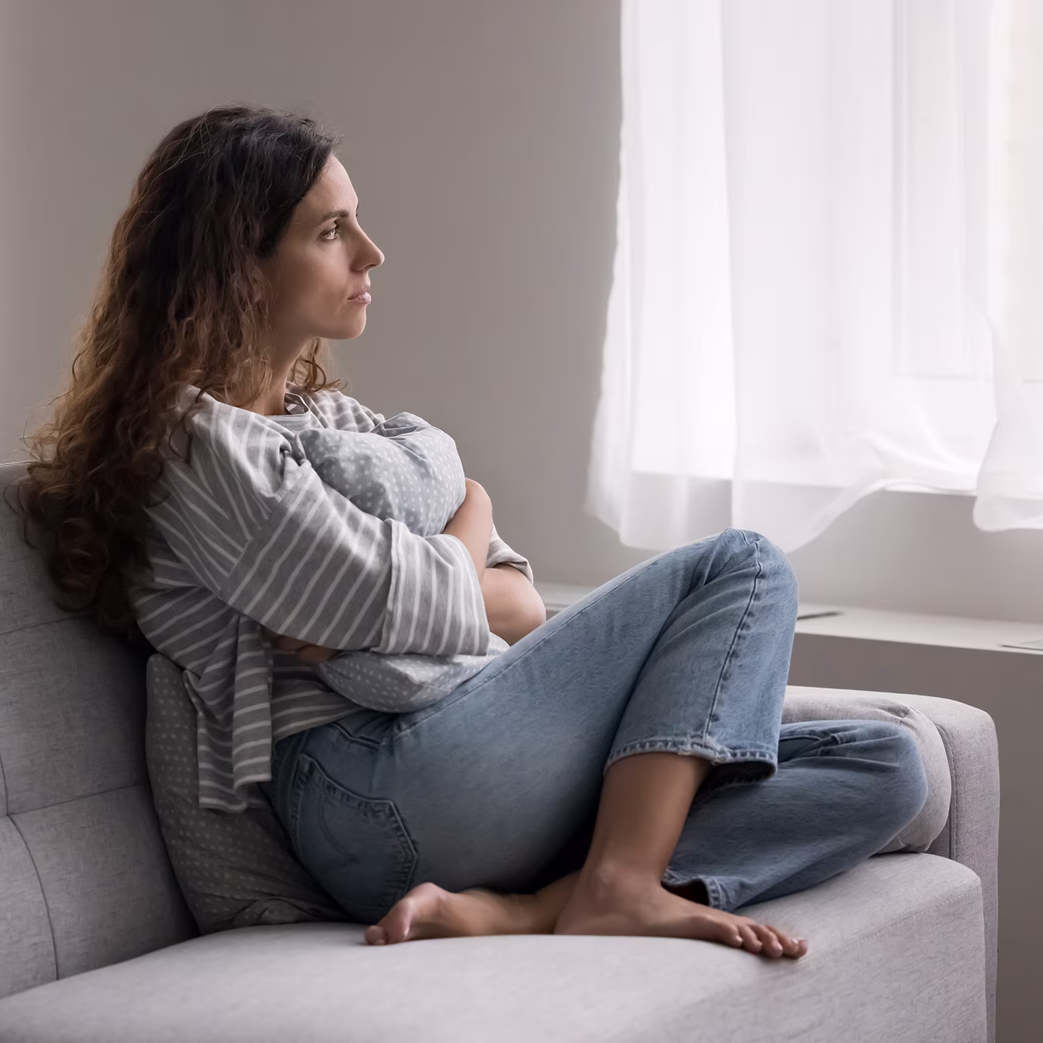 Woman sits curled on a couch by a bright window, appearing reflective and concerned