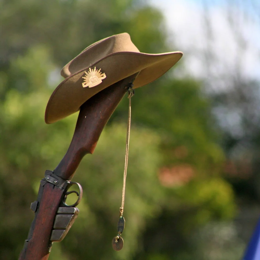 ANZAC Day tribute featuring a vintage Australian Army .303 rifle with slouch hat and rising sun badge, soldier’s dog tags, floral wreath, and the Australian flag in the background
