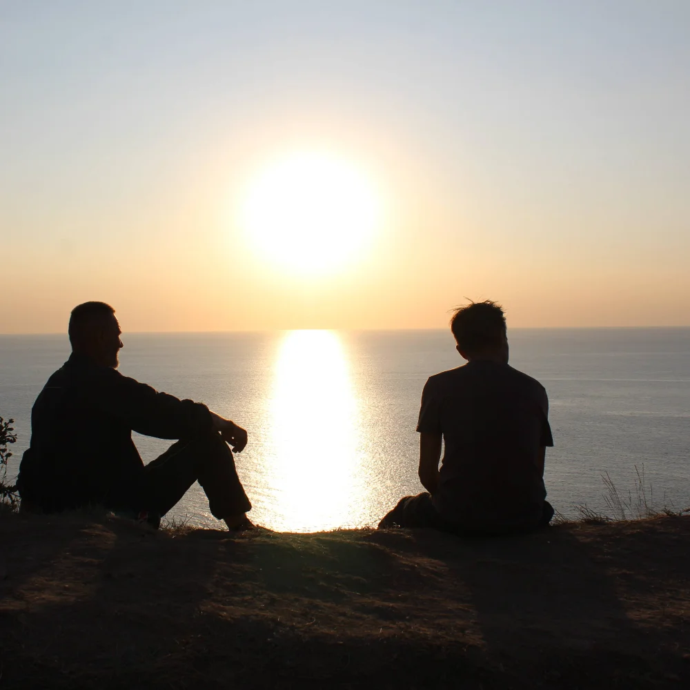 Silhouette of an executive rehab client and their counsellor sitting on the beach at sunset, engaged in a reflective one-on-one conversation.