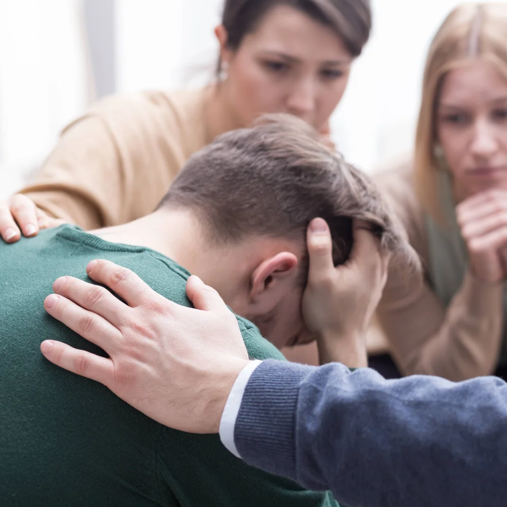 Young man sitting with his head in his hands during a family-led intervention, surrounded by supportive loved ones encouraging him to take the first step toward recovery.