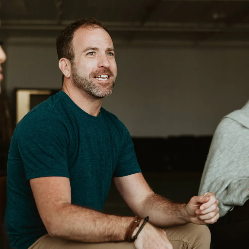 A male resident sits in a group therapy session, with a female resident to his right and a female therapist to his left. He’s speaking to the group.