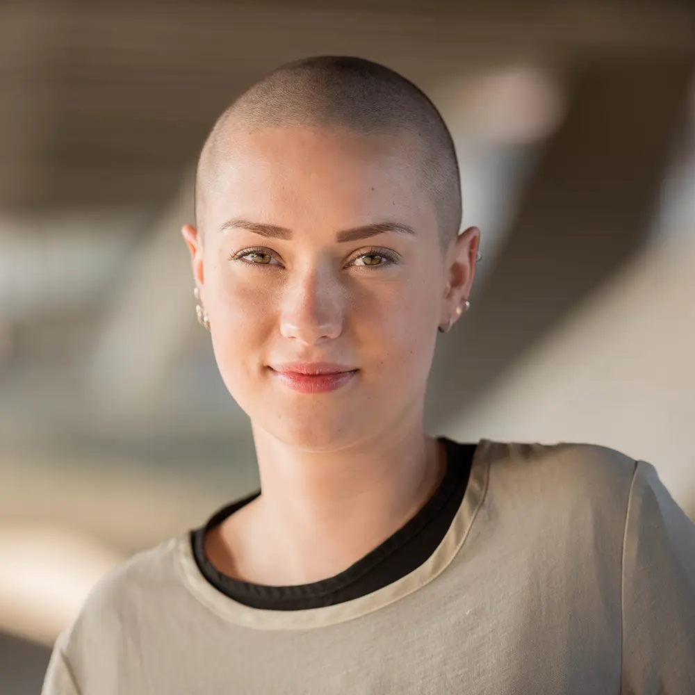 A young woman in her twenties, with a shaved head and multiple ear piercings, smiling at the camera in a candid moment while walking around the rehab centre. If you have private health insurance, it may make residential rehab more accessible.