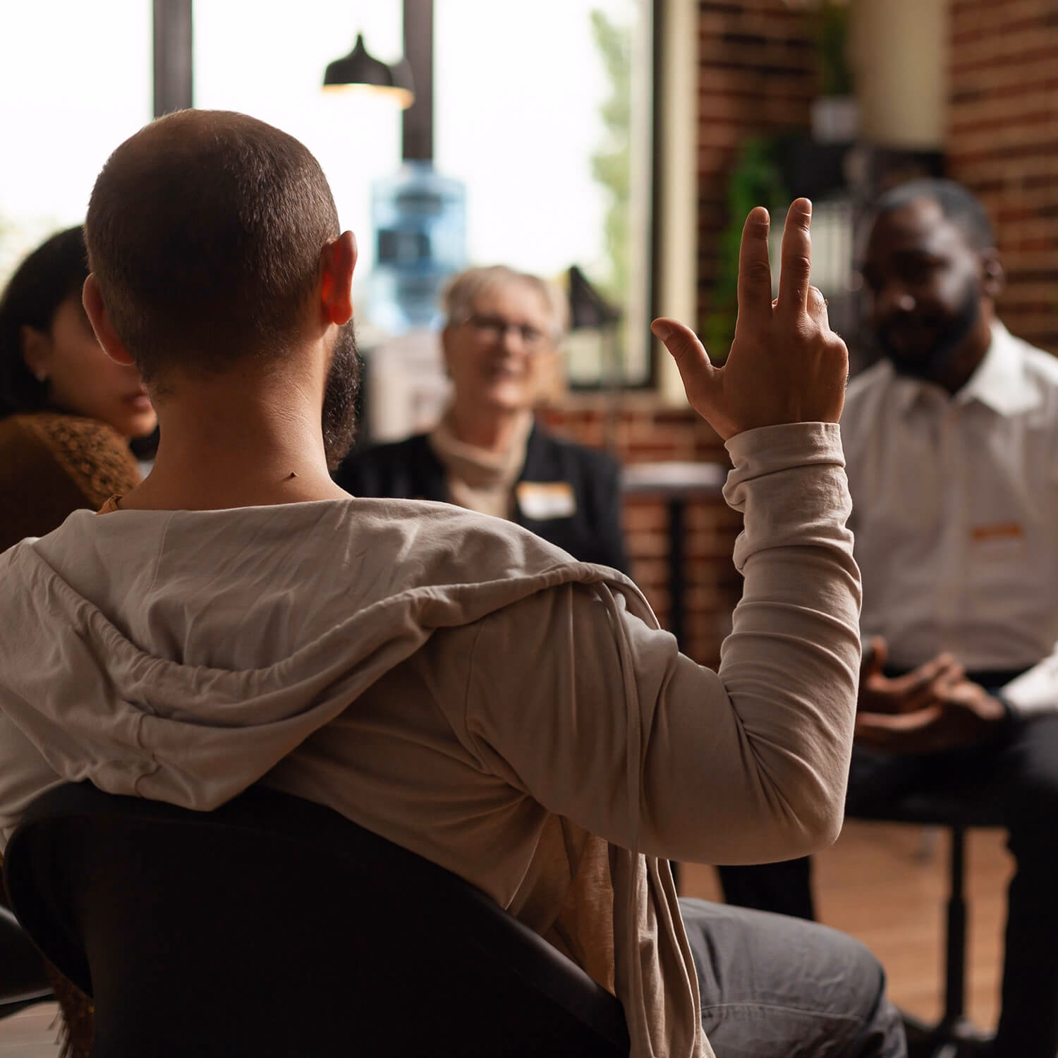 A group therapy session where a man raises his hand to speak while others listen attentively in a bright counselling room — representing open communication, trust, and emotional recovery during rehabilitation.