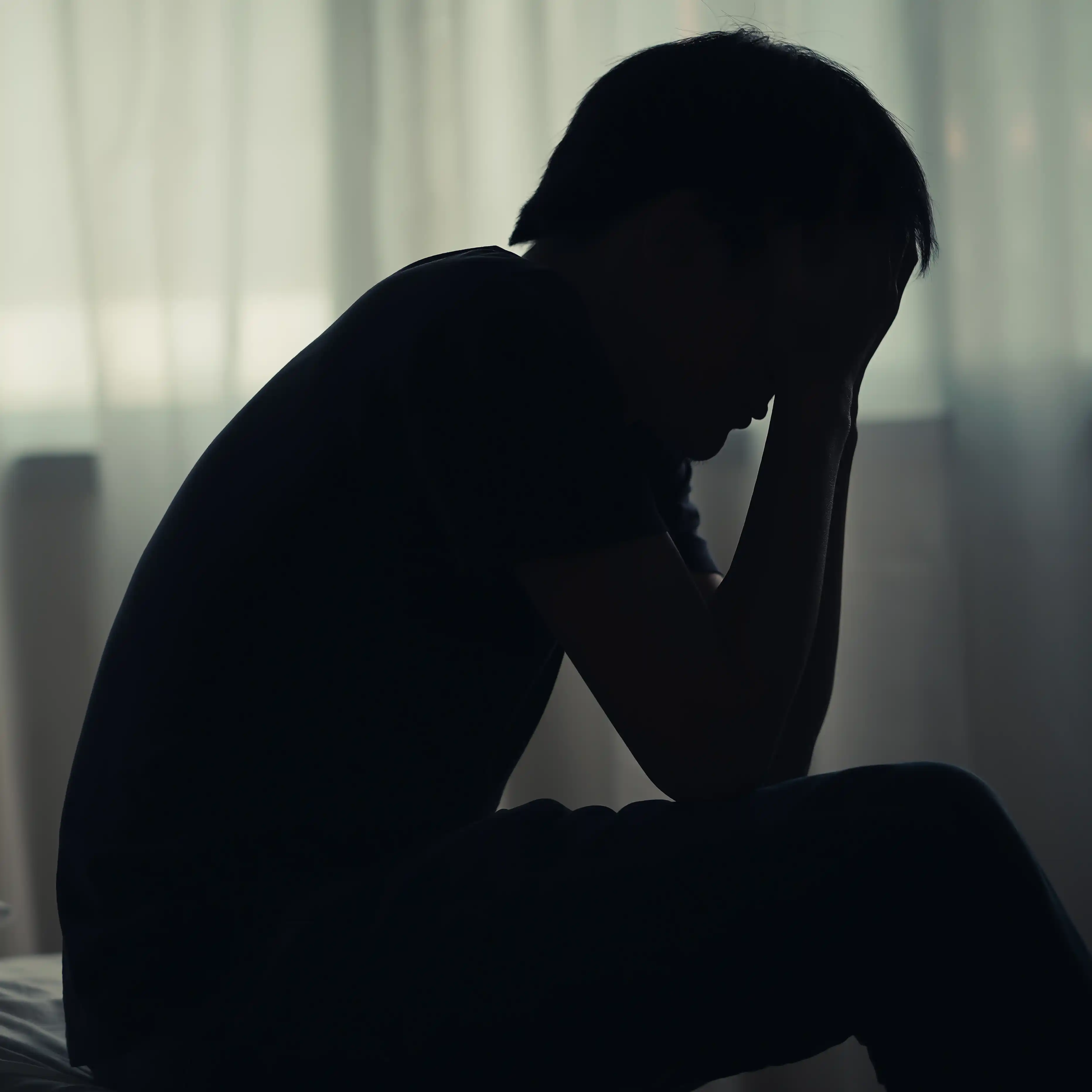 A man sits alone on a bed in a dimly lit room, with his head in his hands, overwhelmed by stress and worry, reflecting the emotional struggles of heroin addiction and the need for heroin rehab support.