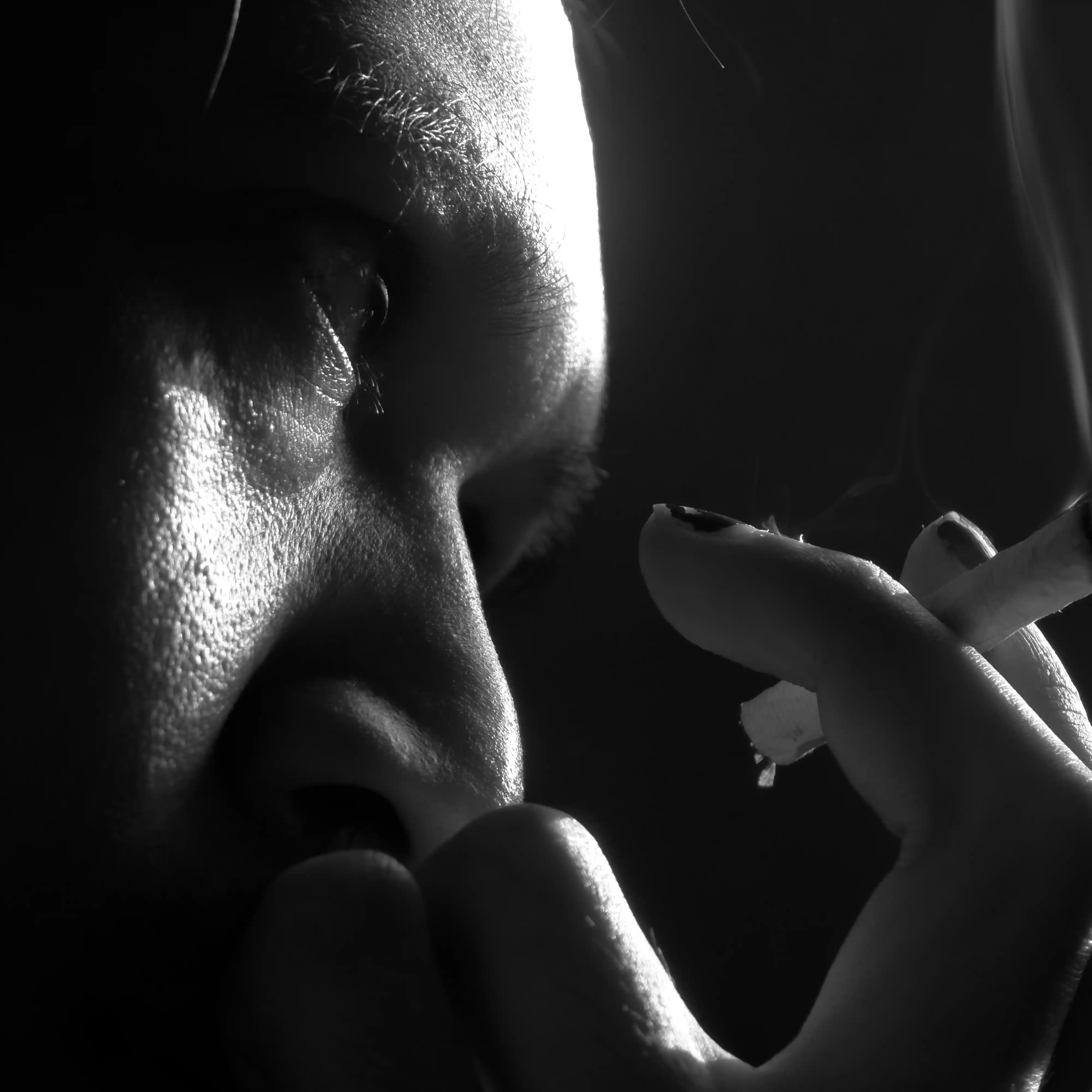 Close-up of a person’s face in dramatic lighting, holding a lit cigarette near their mouth with smoke rising in the dark background.