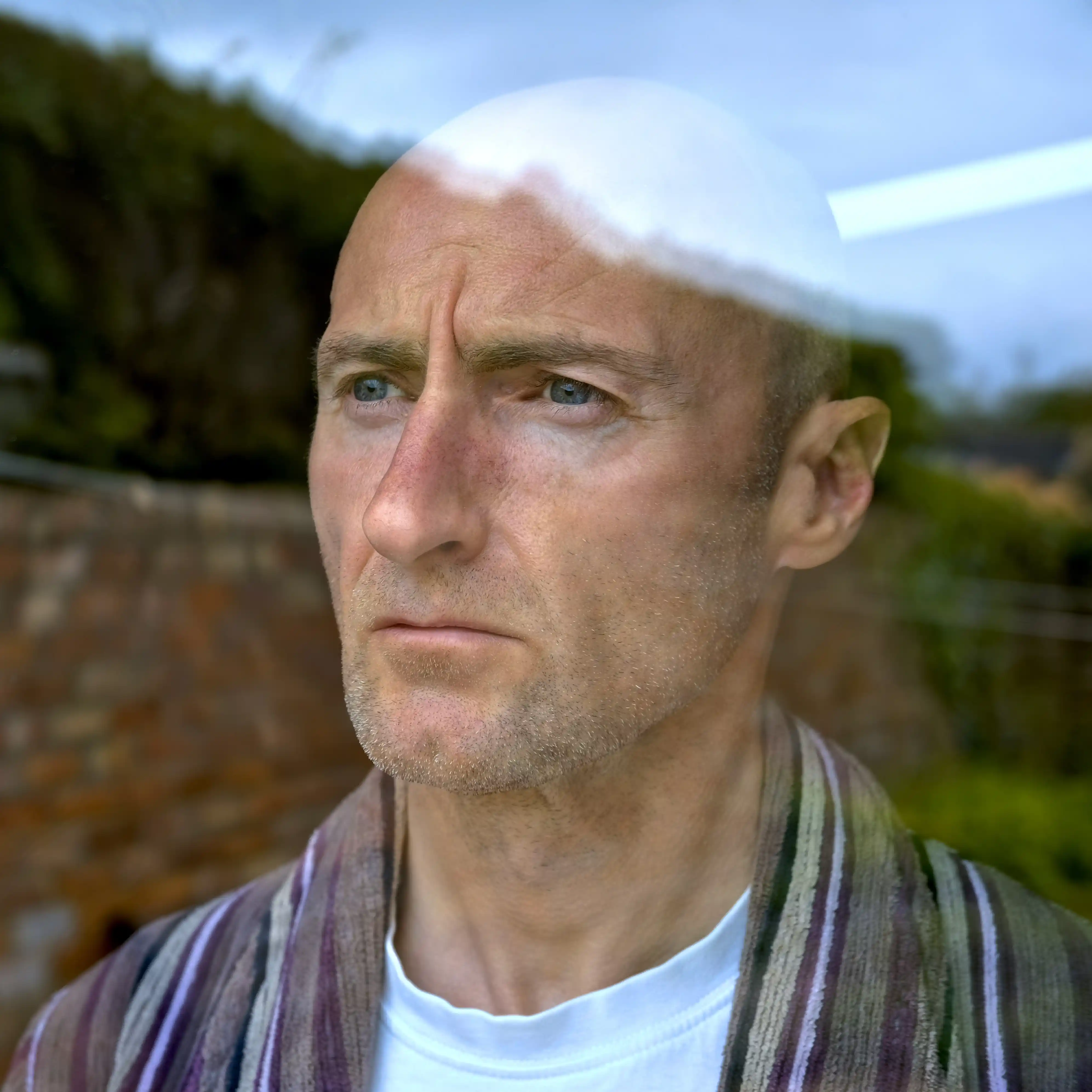 Middle-aged man with a serious expression looking out a window, reflection of brick wall and garden visible, symbolising reflection, stress, or addiction recovery.