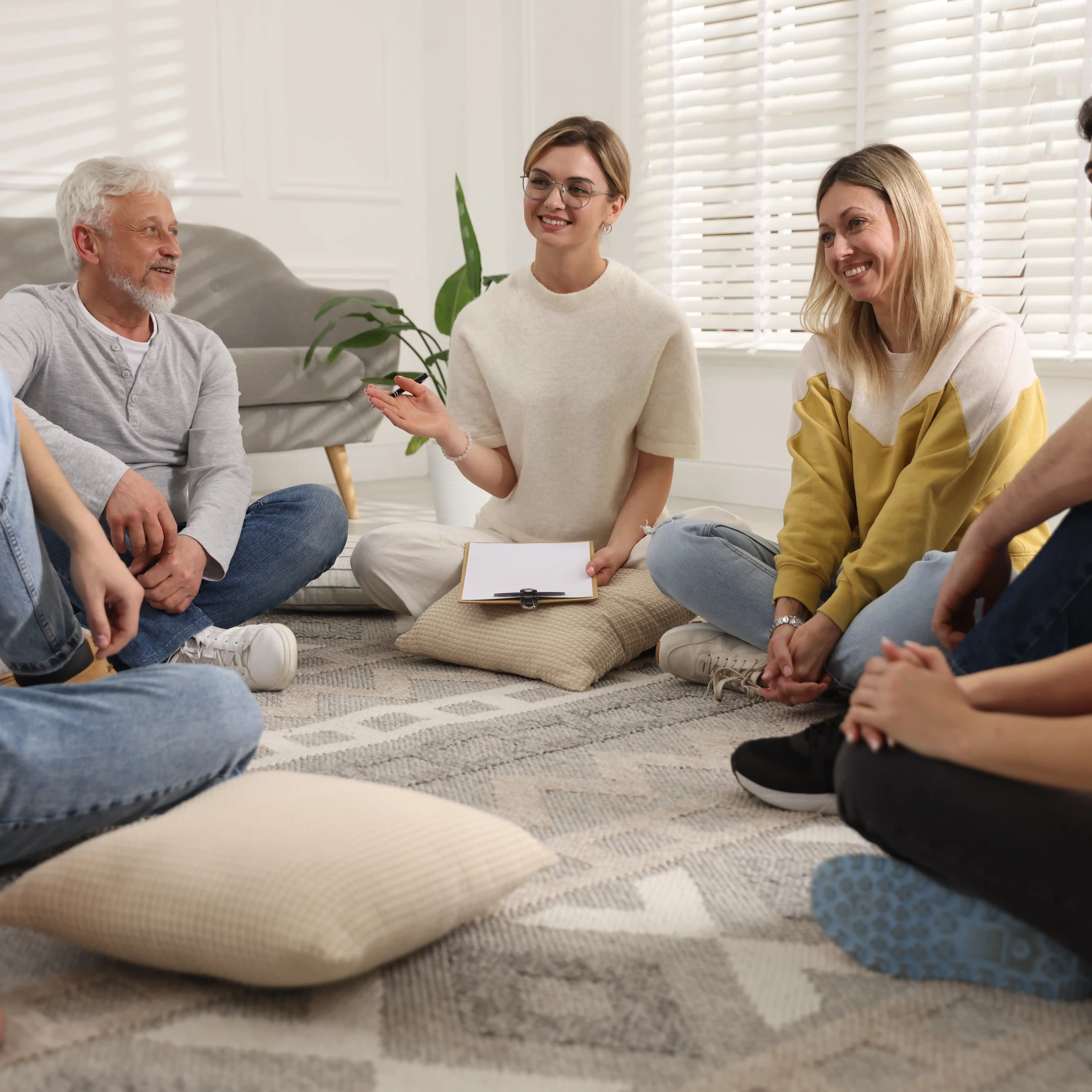 Group of people sitting in a circle on the floor with a therapist leading the discussion, smiling and engaged in a supportive group therapy session.