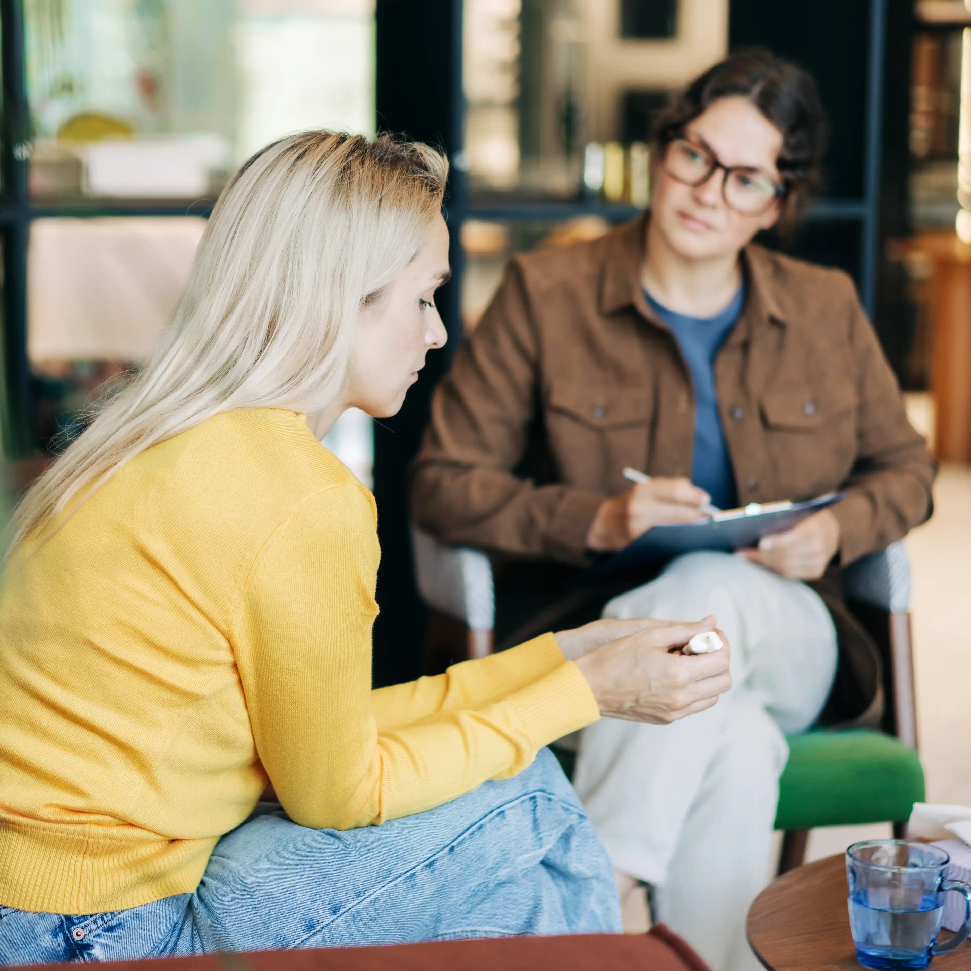A woman in a yellow sweater sits and talks with a therapist holding a clipboard during a counseling session in a comfortable, modern office setting.
