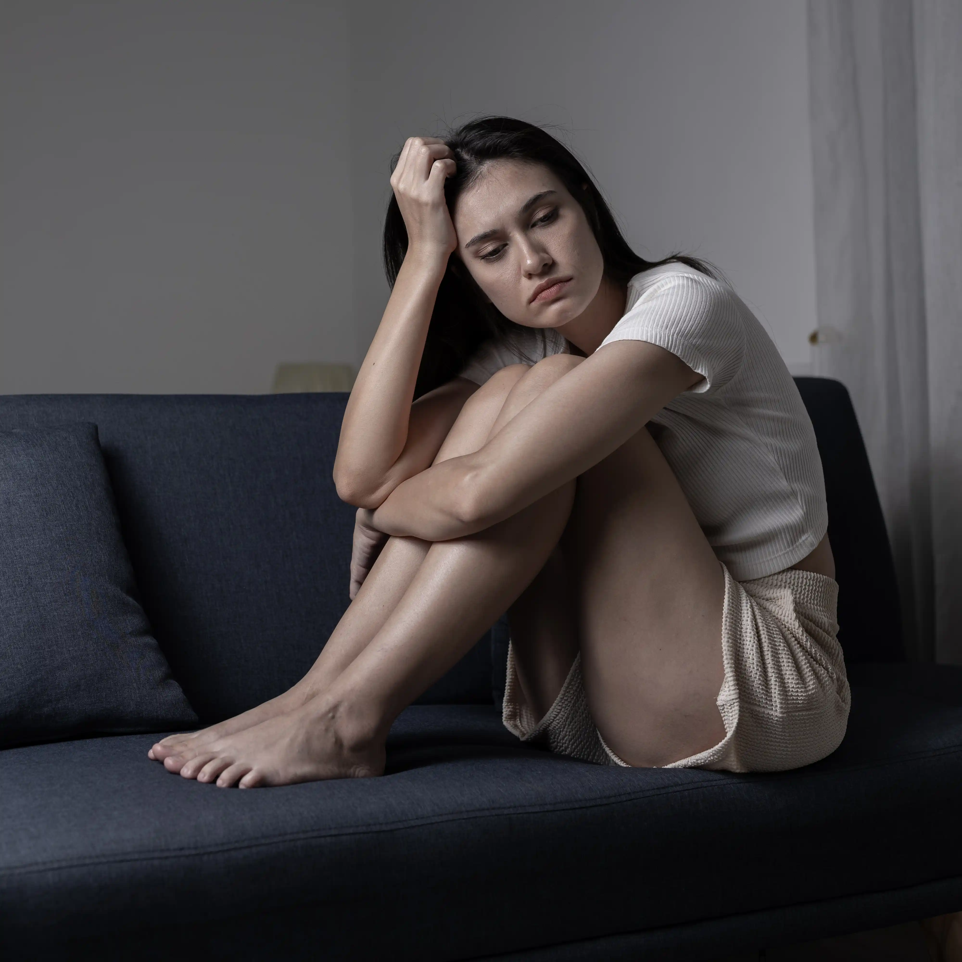 A young woman sits curled up on a dark sofa, resting her head on her hand and looking down with a sad or pensive expression in a dimly lit room.
