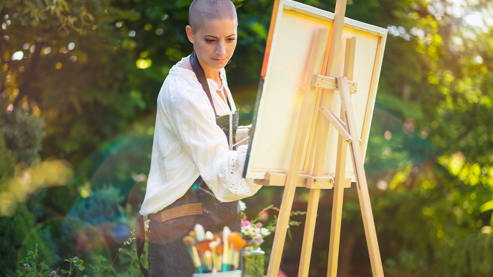 A young woman relaxing while painting an art canvas outdoors in her garden.