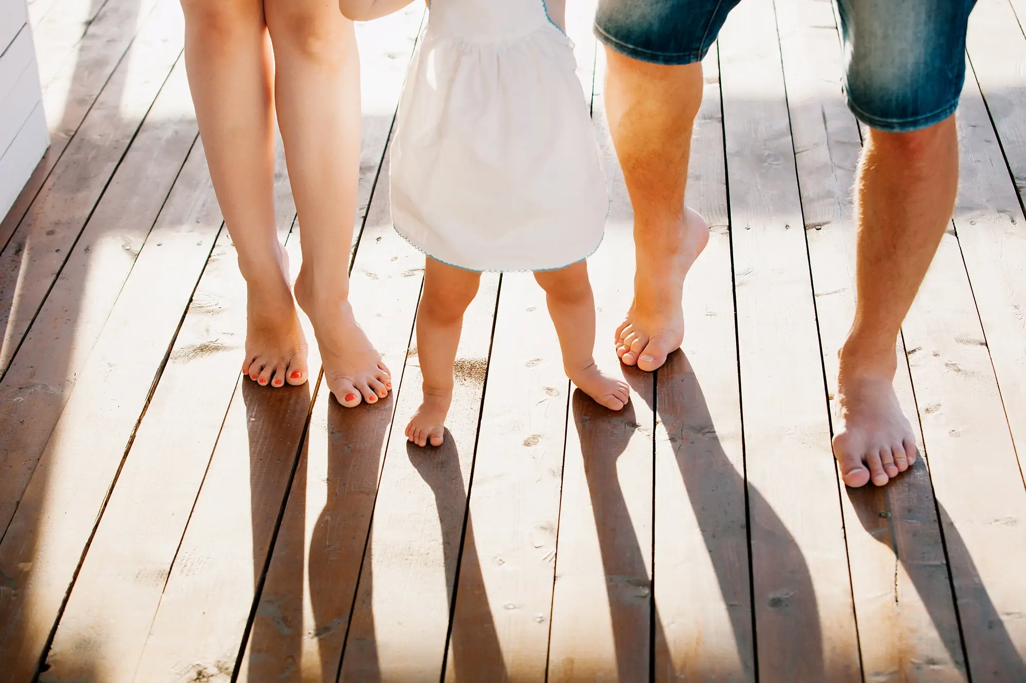 Close-up of a mother, father, and daughter standing barefoot on a wooden floor at sunset, their feet touching as they enjoy a quiet, joyful moment together — reflecting the healing and connection made possible after a successful intervention and recovery journey.