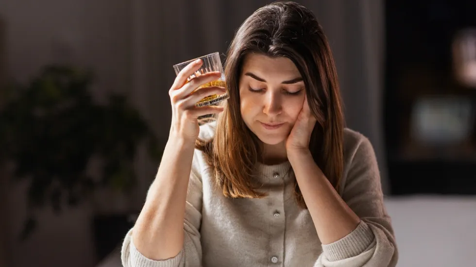 A woman sitting alone in a dimly lit room holding a glass of alcohol to her head, looking down with a troubled expression.