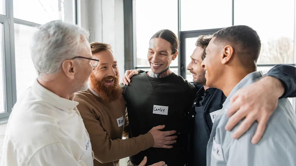 A group of men standing close together in a supportive embrace, smiling and wearing name tags during a recovery or support meeting.