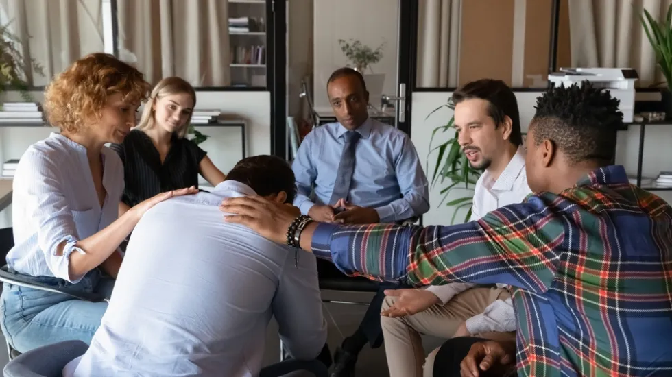 A group of people in a support meeting comforts a distressed man who is leaning forward, with several members placing reassuring hands on his back and shoulders