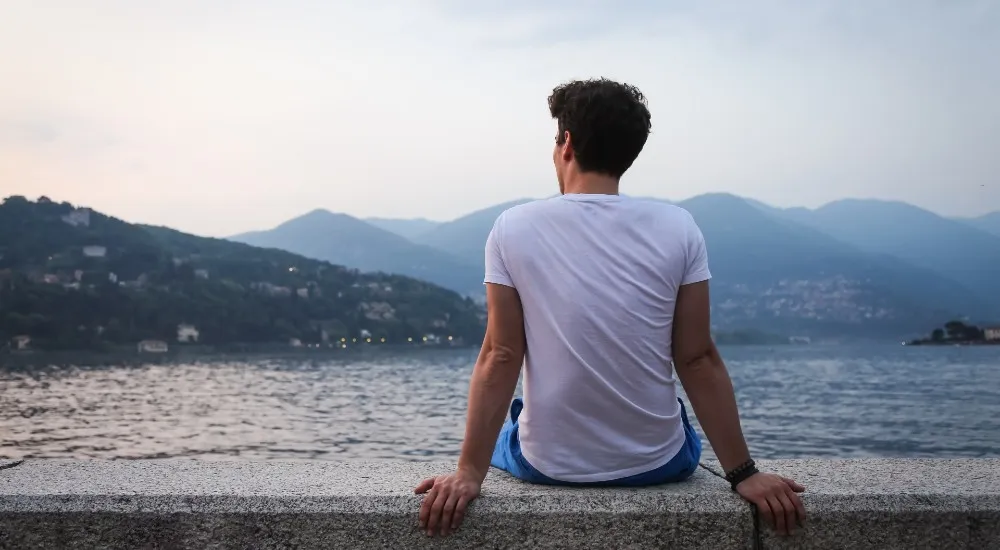A man sits on a stone ledge facing a lake, looking out at the distant mountains and shoreline.