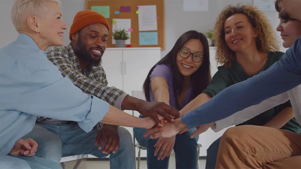 A diverse group of people sitting in a circle and stacking their hands together in the centre, smiling and showing unity and support.