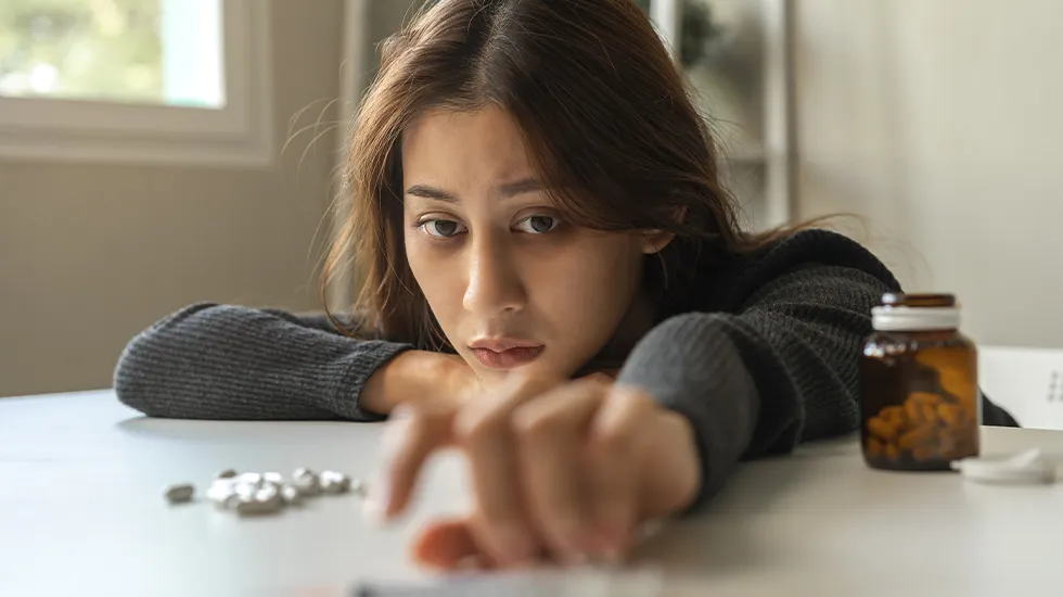 A young woman leaning over a table with a distressed expression, reaching toward scattered pills beside an open pill bottle.