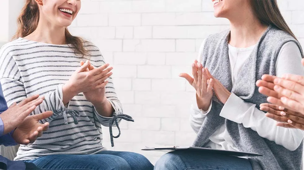 People seated in a support group circle, smiling and clapping as they encourage someone speaking.