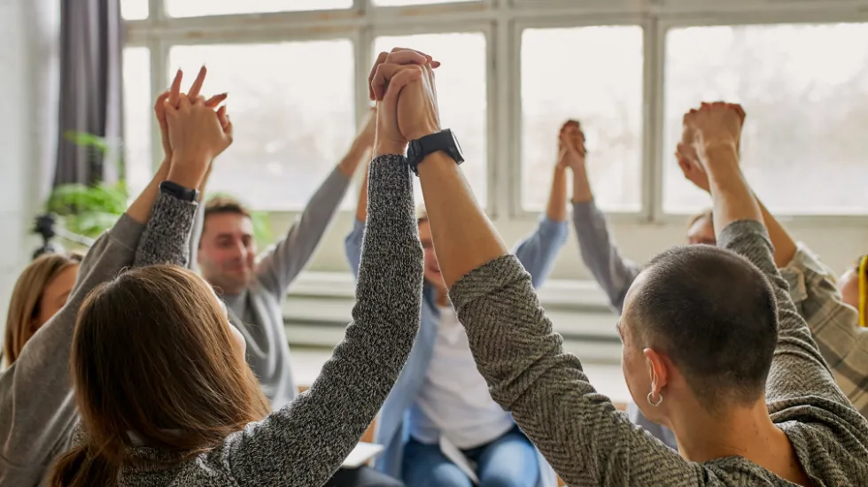 A group of people sitting in a circle, raising their joined hands high in celebration or unity during a support or group therapy session.