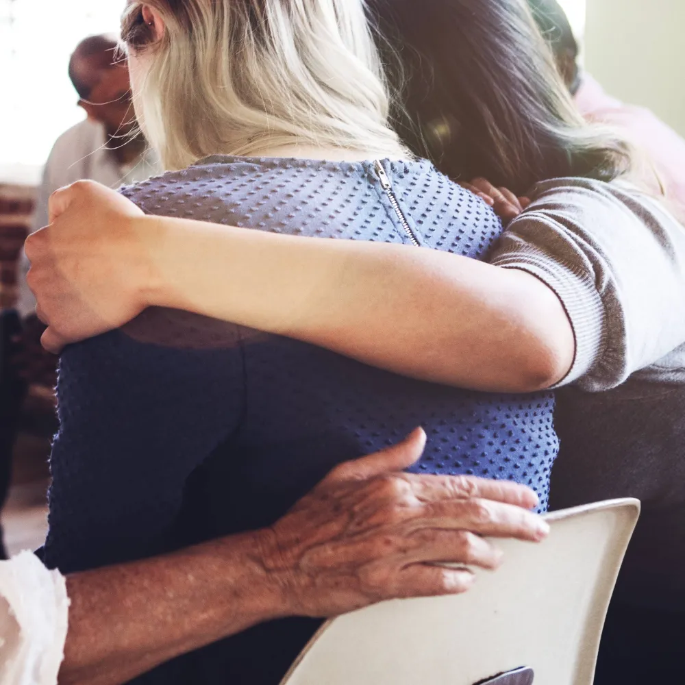 A therapy session, where a few of the residents have their arms over the shoulders of a female resident who’s sharing her story. 