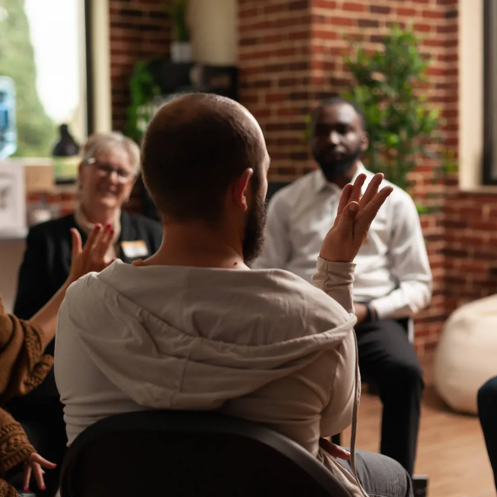 A group therapy session with male and female residents sitting in a circle, joined by a therapist. The faces are out of focus. 