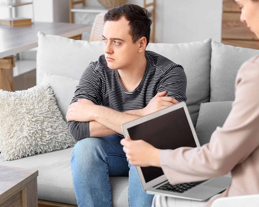 A therapist, during a session, with her laptop sitting beside a man on a sofa.
