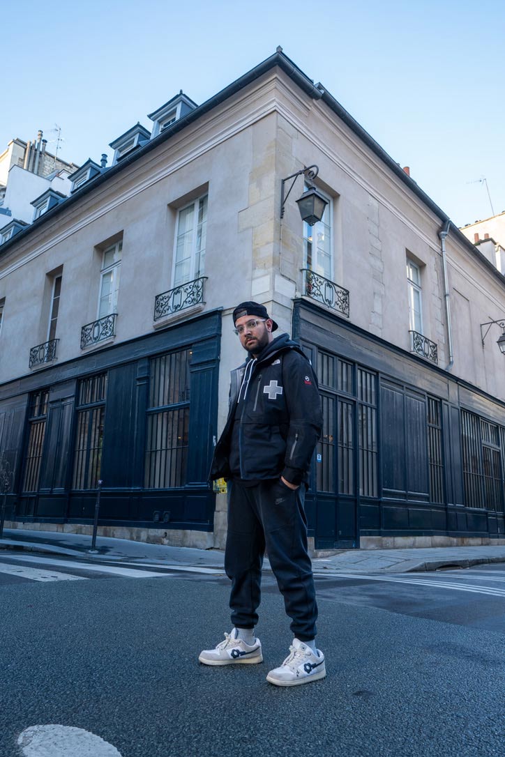 Ishaan Mishra Man wearing a black jacket, joggers, and white sneakers standing on a city street corner in front of a beige and dark blue building with ornate window railings.