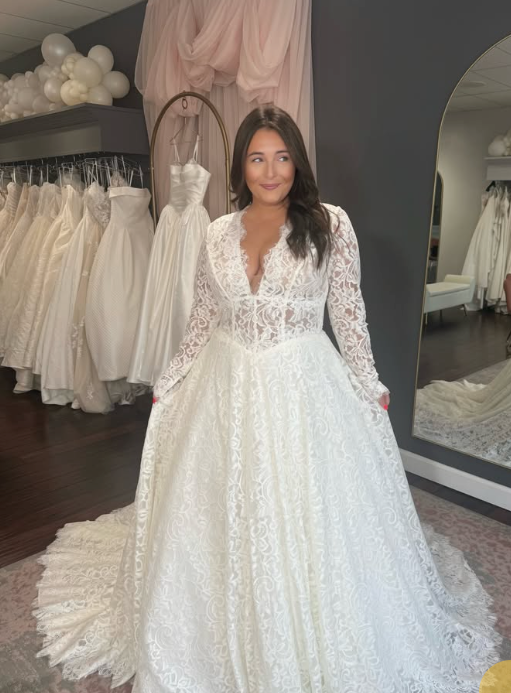 Model in a long-sleeve lace A-line wedding dress with a plunging V-neck and sheer bodice in a bridal shop