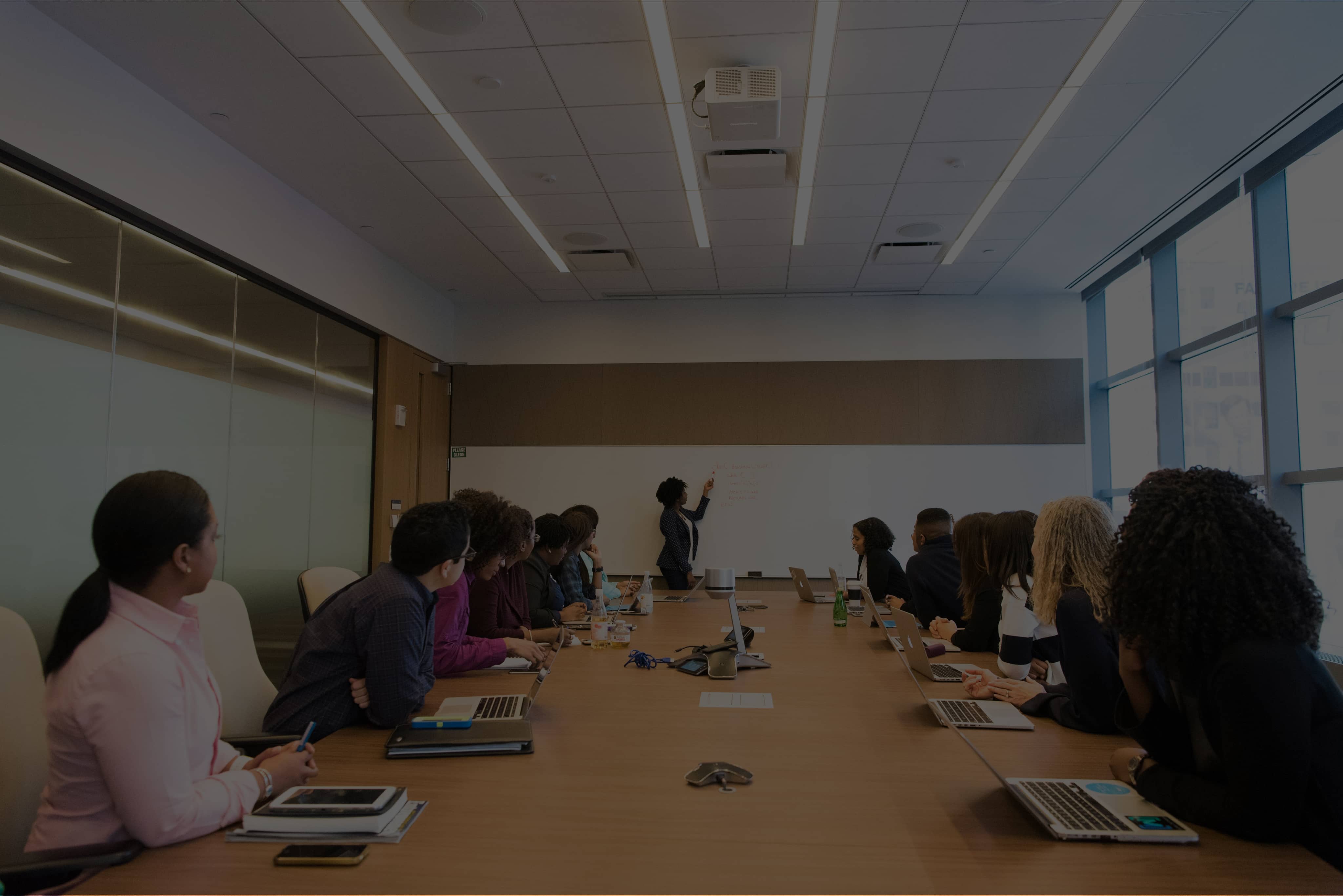 A diverse group of people seated around a long conference table, attentively listening to a woman standing and writing on a whiteboard in a bright meeting room.
