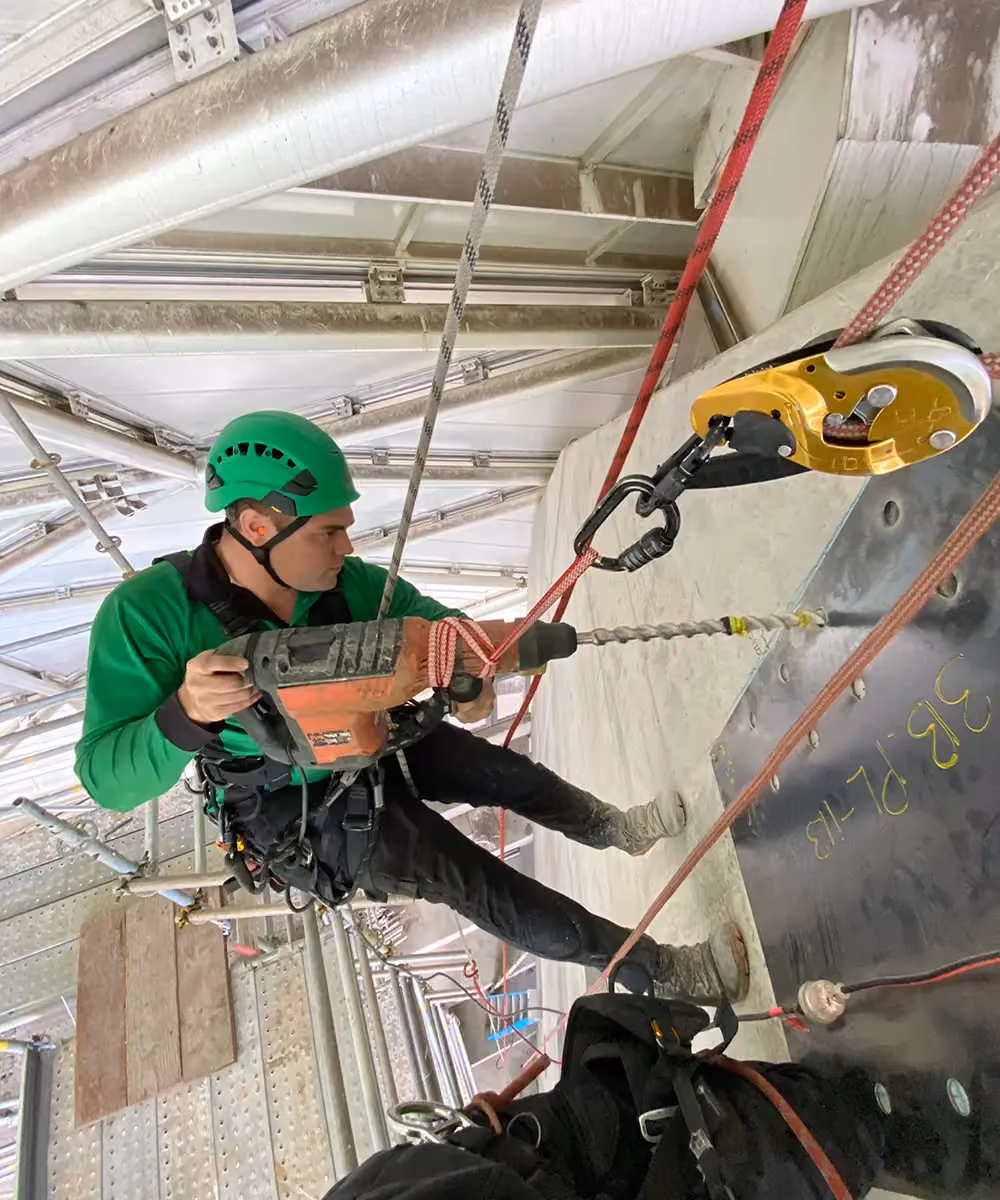 A Bax rope access specialist, suspended above a multistorey gap, using a power drill to create an anchor point in a concrete wall.