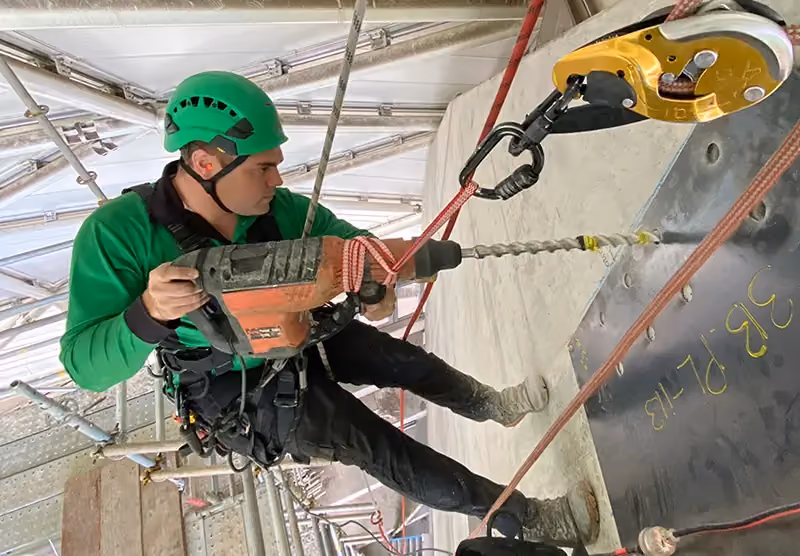 A Bax rope access specialist, suspended above a multistorey gap, using a power drill to create an anchor point in a concrete wall