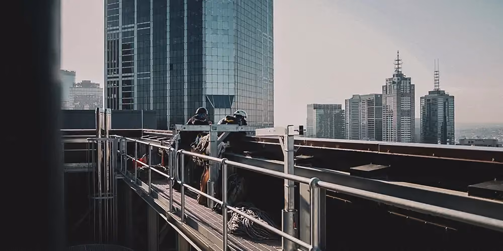 Two rope access technicians working from a rooftop gantry platform with safety harnesses, positioned near the ME Bank building in Melbourne.