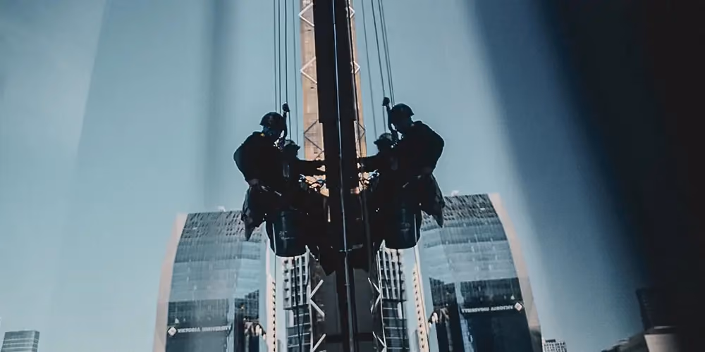 Rope access technician abseiling a city skyscraper with a full gear setup, captured with a mirrored reflection on the building’s glass surface.