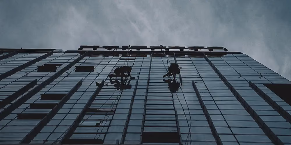 Silhouetted rope access technicians suspended mid-air while cleaning or inspecting a high-rise glass façade.