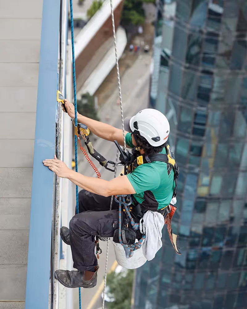 A high-rise cleaner in a safety harness and helmet is suspended on ropes.