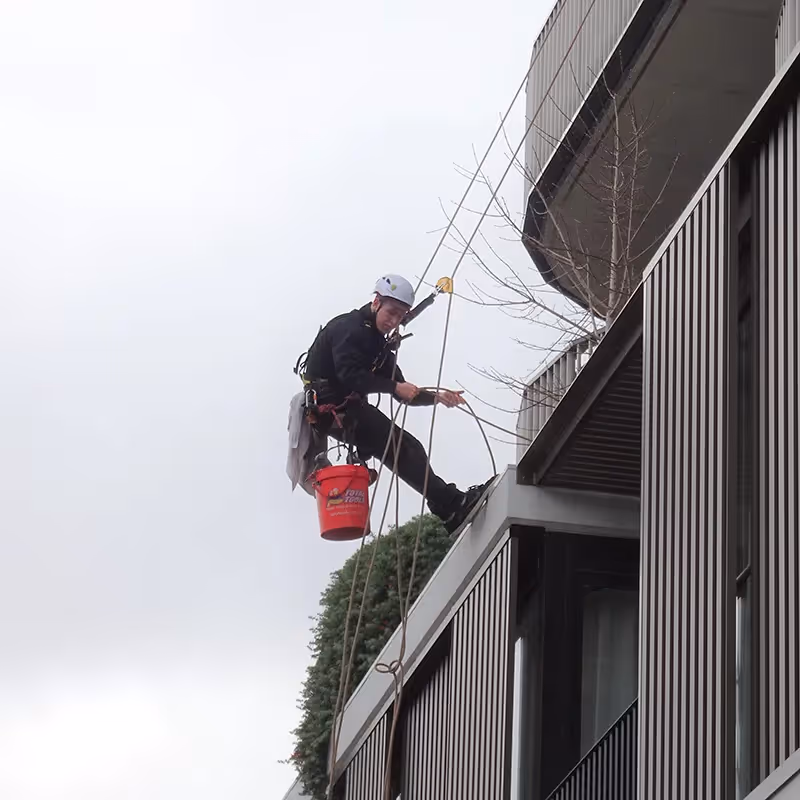 A rope-access technician wearing a helmet and harness is suspended over the edge of a modern building.