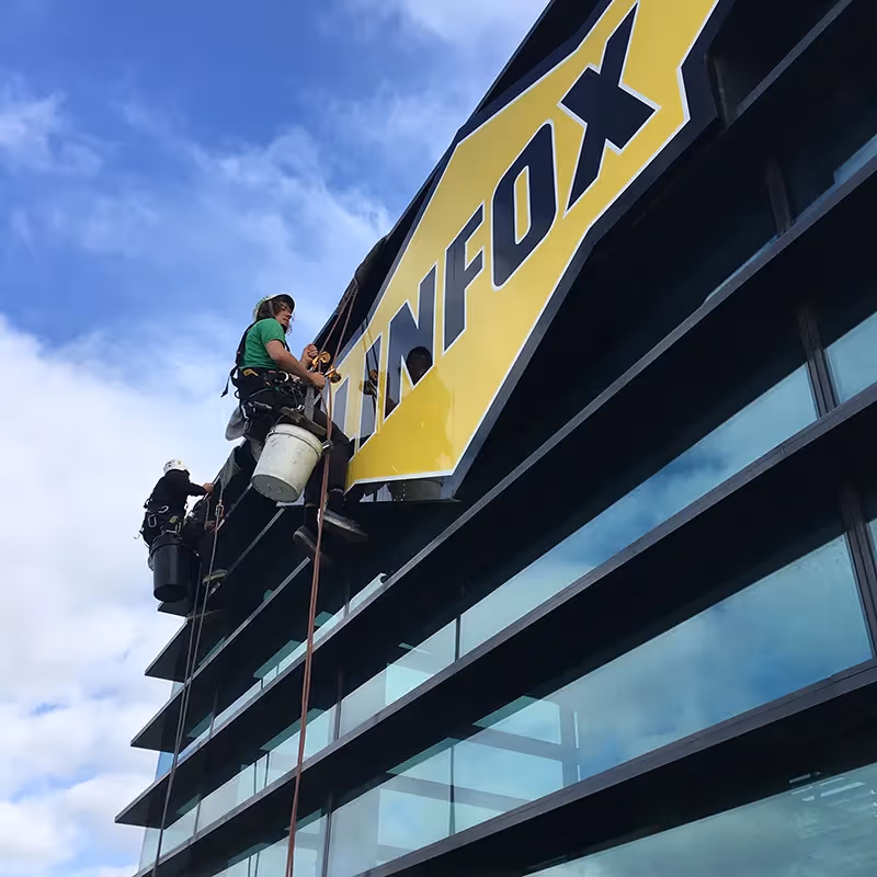 Two high-rise rope-access technicians in harnesses work on the side of a tall building.