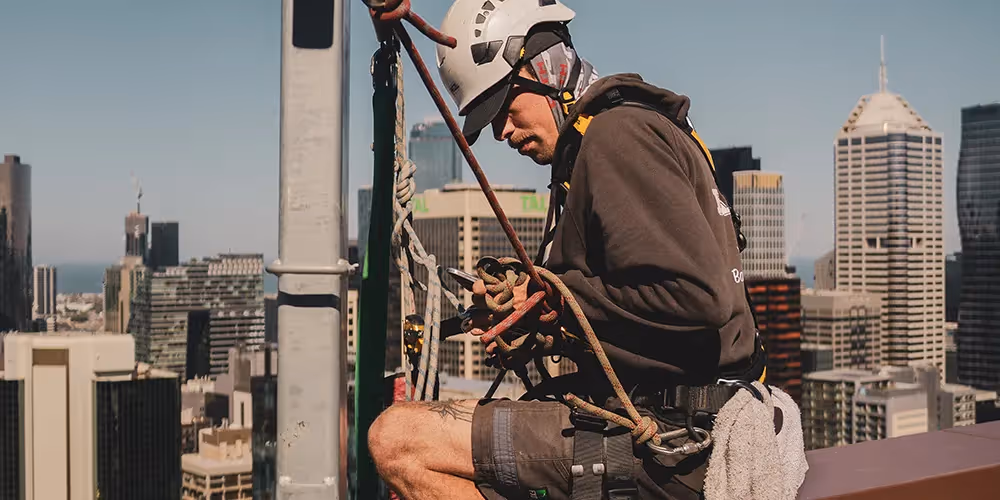 Rope access technician securing safety lines to an anchor point on a high-rise rooftop, with the Melbourne skyline in the background.