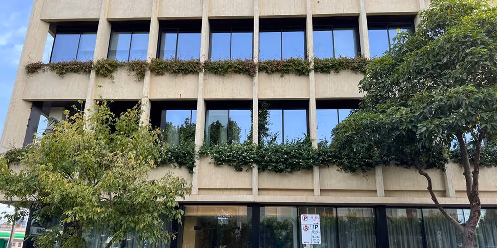 Three-storey commercial building with vertical gardens and greenery maintained on upper-level ledges and window boxes, surrounded by street trees.