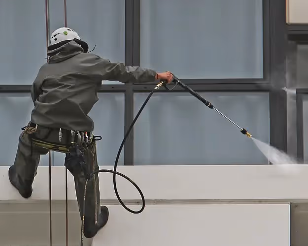 A rope-access worker in safety gear and a helmet pressure washes the exterior of a high-rise building. 