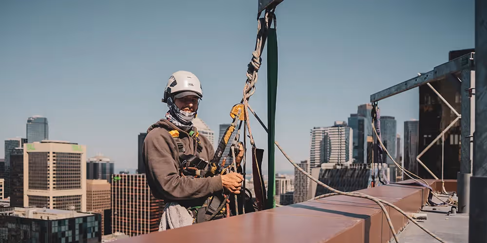 Rope access technician harnessed and ready for high-rise work, standing at a rooftop anchor point with Melbourne city skyline in the background.