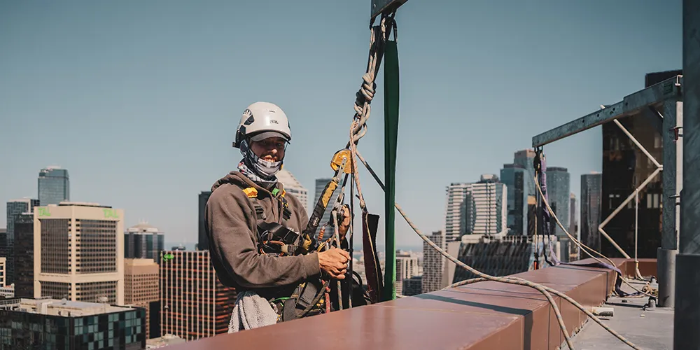 Rope access technician harnessed and ready for high-rise work, standing at a rooftop anchor point with Melbourne city skyline in the background.