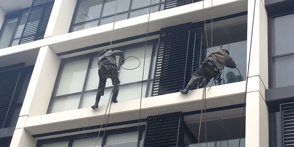 Two rope access technicians using pressure hoses while suspended on ropes, cleaning the exterior windows and facade of a multi-storey building.