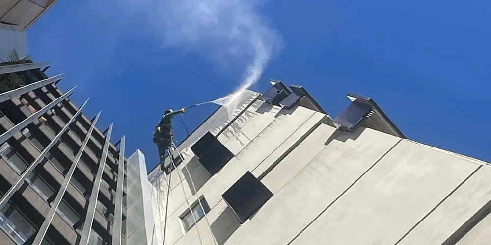 Rope access worker using high-pressure water spray to clean a tall building’s exterior under a clear blue sky.